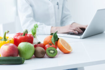na zdjęciu na biurku stoi miska warzyw i owoców a za biurkiem widać lekarza w białym fartuchu